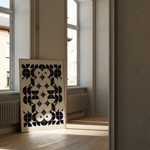 May include: A black and white geometric print in a gold frame hangs on a white wall in a room with large windows. The print features a repeating pattern of circles, semi-circles, and squares.