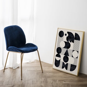 Puede incluir: Una silla de terciopelo azul con patas doradas se encuentra frente a una pared blanca con una impresi&oacute;n geom&eacute;trica en blanco y negro enmarcada. La impresi&oacute;n presenta c&iacute;rculos, cuadrados y rayas.