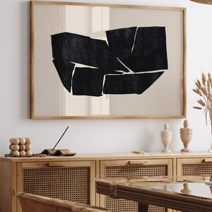 K&ouml;nnte beinhalten: Ein gerahmter abstrakter Kunstdruck mit einem geometrischen Design in Schwarzwei&szlig;. Der Druck h&auml;ngt an einer wei&szlig;en Wand &uuml;ber einem Holzschr&auml;nkchen mit geflochtenen Rattan-T&uuml;ren.