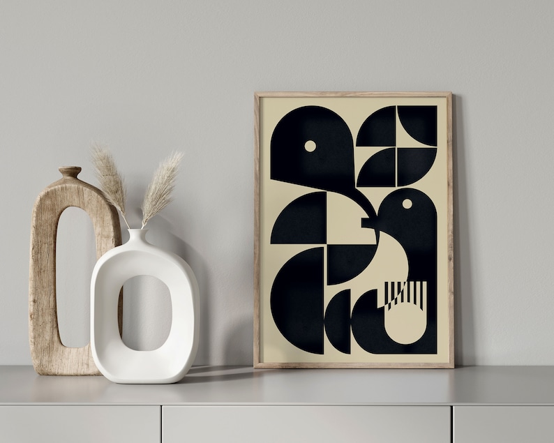 May include: A framed print featuring a geometric design in black and white. The design depicts a bird-like shape with a white dot in the centre of its head. The background is a light beige colour.