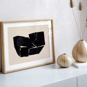 K&ouml;nnte beinhalten: Ein gerahmter abstrakter Kunstdruck mit einem schwarzen geometrischen Design auf beigefarbenem Hintergrund. Der Druck ist in einem hellen Holzrahmen und liegt auf einer hellgrauen Oberfl&auml;che mit zwei Holzvasen und einem Buch.