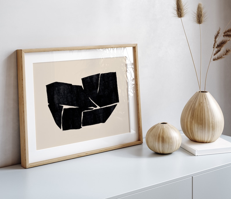 K&ouml;nnte beinhalten: Ein gerahmter abstrakter Kunstdruck mit einem schwarzen geometrischen Design auf beigefarbenem Hintergrund. Der Druck ist in einem hellen Holzrahmen und liegt auf einer hellgrauen Oberfl&auml;che mit zwei Holzvasen und einem Buch.