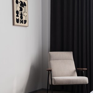 Puede incluir: Un sill&oacute;n beige con patas de metal negro y apoyabrazos de madera se encuentra en una habitaci&oacute;n con una pared blanca y cortinas negras. Un estampado enmarcado con un dise&ntilde;o geom&eacute;trico en blanco y negro cuelga de la pared.