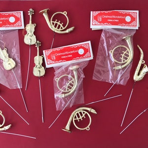 May include: Gold-colored Christmas tree ornaments in the shape of musical instruments, including violins, trumpets, French horns, and a saxophone. Some ornaments are packaged with red and white labels that read "Christmas Wonderland Trim A Tree."