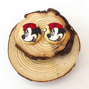 May include: Two yellow fabric buttons with a cartoon image of Minnie Mouse. The buttons have red bows and black eyes. The buttons are on a wooden surface.