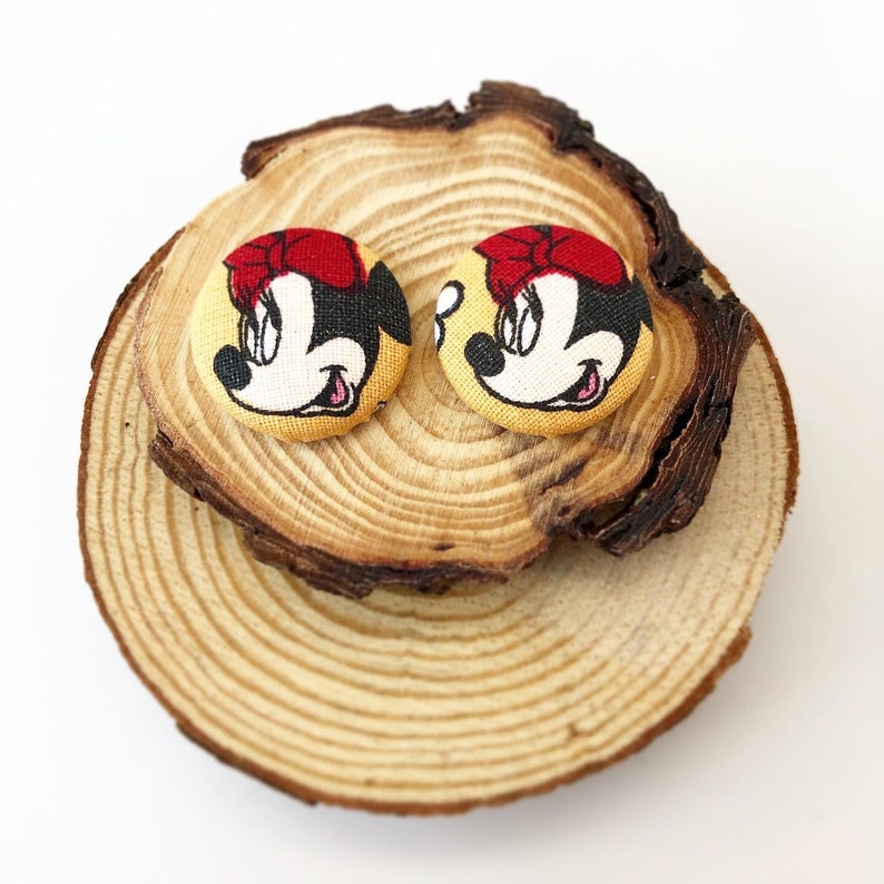May include: Two yellow fabric buttons with a cartoon image of Minnie Mouse. The buttons have red bows and black eyes. The buttons are on a wooden surface.