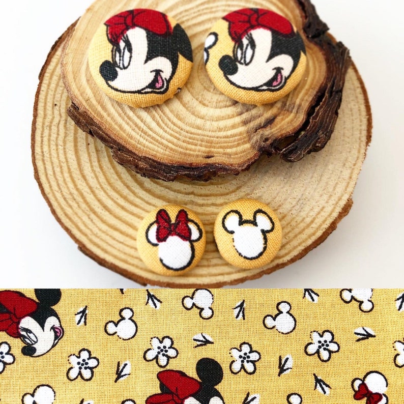 May include: Four fabric buttons featuring Mickey Mouse characters. Two buttons have a red bow and a white face with black eyes and a black nose. Two buttons have a white face with black eyes and a black nose. The buttons are on a wooden surface.