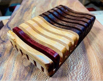 May include: A wooden soap dish with a wavy design. The dish is made of three different types of wood: light wood, dark wood, and a reddish-purple wood.