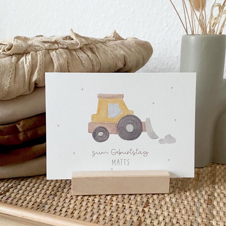 Puede incluir: Una tarjeta de felicitaci&oacute;n rectangular blanca con una ilustraci&oacute;n en acuarela de una excavadora de construcci&oacute;n amarilla y marr&oacute;n con una pala gris. El texto "zum Geburtstag" y "MATTS" est&aacute; impreso en gris oscuro debajo.