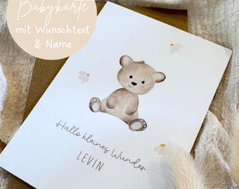 Könnte beinhalten: Eine weiße, rechteckige Grußkarte mit einer Aquarellillustration eines sitzenden braunen Teddybären, umgeben von kleinen weißen Blumen. Die Karte zeigt den Text "Hallo kleines Wunder" und den Namen "LEVIN" in schwarzer Schrift.