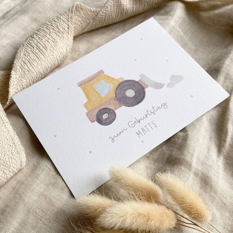 Puede incluir: Una tarjeta de felicitaci&oacute;n rectangular blanca con una ilustraci&oacute;n en acuarela de un veh&iacute;culo de construcci&oacute;n amarillo con ruedas de color gris oscuro. La tarjeta incluye el texto "zum Geburtstag MATTS" en una fuente gris simple.