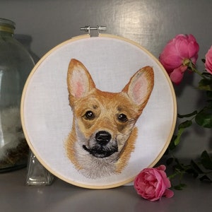 May include: Embroidered portrait of a corgi dog on white fabric stretched within a wooden embroidery hoop. The dog has brown and tan fur with a black nose and a slight smile. The background is a neutral gray.