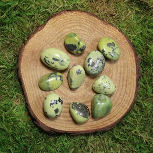 May include: A collection of nine green and black serpentine tumbled stones on a wooden surface. The stones are smooth and polished, with a variety of patterns and textures.