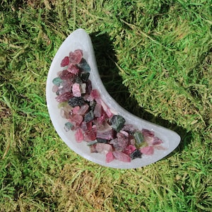 May include: A white crescent moon shaped dish filled with a variety of pink, green, and black gemstones. The dish is sitting on a bed of green moss.