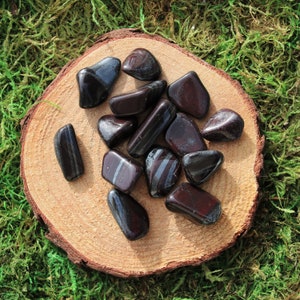 May include: A collection of black and silver hematite tumbled stones on a wooden surface. The stones are smooth and polished, with a natural, earthy look.