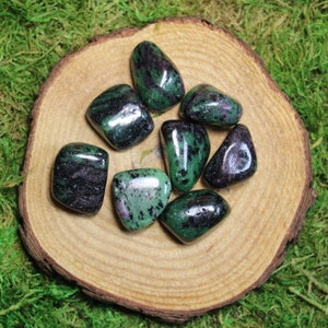 May include: A collection of seven green, black, and purple tumbled stones. The stones are smooth and polished, and they are arranged in a circle on a wooden surface.