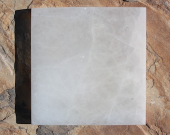 May include: A white square-shaped piece of selenite crystal resting on a brown, textured surface.