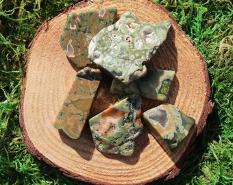 May include: A collection of six green, brown, and white moss agate stones on a wooden slice. The stones are irregularly shaped and have a variety of patterns.