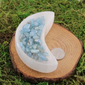 May include: A white selenite crescent moon shaped dish filled with light blue aquamarine chips. The dish is sitting on a wooden surface.
