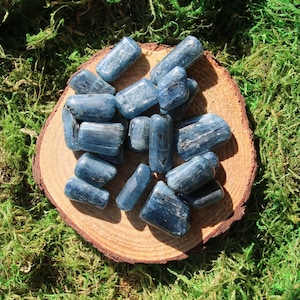 May include: A collection of blue kyanite tumbled stones on a wooden slice. The stones are smooth and polished, with a light blue color and a slightly iridescent sheen.