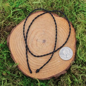 May include: A black braided cord necklace, approximately the size of a US quarter, displayed on a wooden surface.