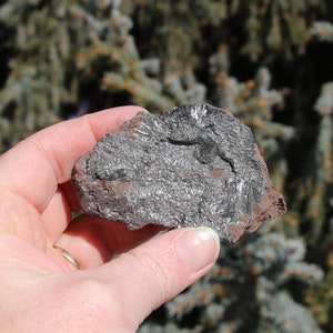 May include: A close-up of a piece of raw, silver-grey, metallic rock with a rough, textured surface. The rock has a reddish-brown hue in some areas.