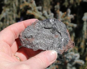May include: A close-up of a piece of raw, silver-grey, metallic rock with a rough, textured surface. The rock has a reddish-brown hue in some areas.