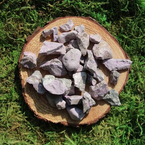 May include: A collection of rough, lilac-colored stones scattered on a round, natural wood slice. The wood slice has a brown bark edge and sits atop a bed of green moss. The stones vary in size and shape, with a mix of light and dark tones.
