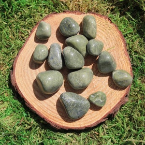 May include: A collection of green serpentine tumbled stones on a wooden slice. The stones are smooth and polished, with a variety of shades of green.