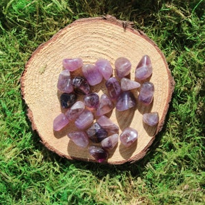May include: A collection of amethyst tumbled stones in various shades of purple, arranged on a wooden slice. The stones are smooth and polished, with a natural, earthy look.