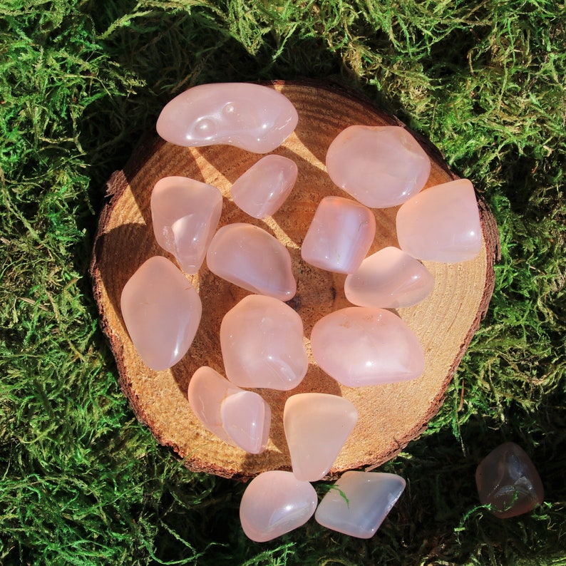 以下が含まれることがあります： 木製の円盤の上に、滑らかで磨かれた表面を持つ、タンブル加工されたローズクォーツの石のコレクションが置かれています。石はピンクと白の様々な色合いで、半透明な外観をしています。