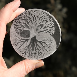May include: A circular, gray stone coaster with a detailed etching of a tree of life design. The tree branches out from the center and has roots that spread out to the edges of the coaster.