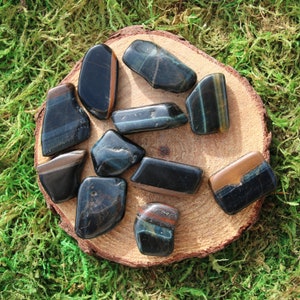 May include: A collection of ten blue tiger's eye tumbled stones on a wooden surface. The stones are various shades of blue and brown with a shimmery effect.