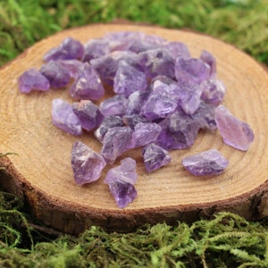 May include: A collection of raw amethyst crystals in various shapes and sizes. The crystals are a deep purple color and are arranged on a round, wooden slice. The background features green moss.