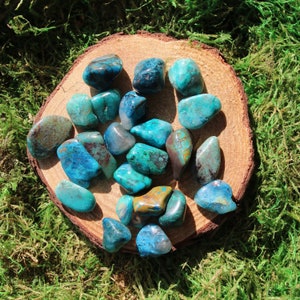 May include: A collection of tumbled blue and green chrysocolla stones on a wooden surface. The stones are various sizes and shapes, with some having a more opaque appearance than others.