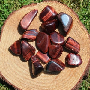 May include: A collection of red tiger's eye tumbled stones, with some showing a silver sheen, on a wooden surface.