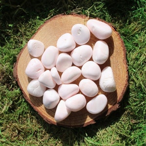 May include: A collection of pink selenite tumbled stones on a wooden surface. The stones are smooth and polished, with a soft, milky white color.