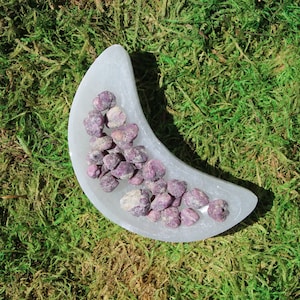 May include: A white crescent moon-shaped dish filled with small, rough, purple and pink ruby crystals.
