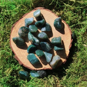 May include: A collection of green moss agate tumbled stones on a wooden slice. The stones are various shades of green and have a natural, polished finish.