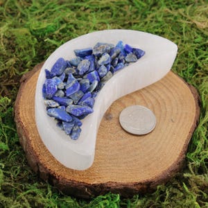 May include: A white selenite crescent moon charging plate filled with blue lapis lazuli chips. The plate is sitting on a wooden base.