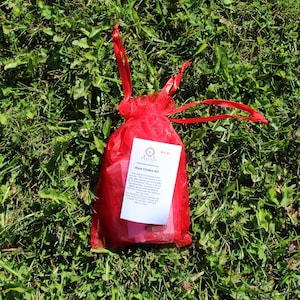 May include: A red mesh bag with a satin ribbon drawstring sits on green grass. A white tag with the text "Root Chakra Kit" is attached to the bag. The bag contains items for spiritual wellness.