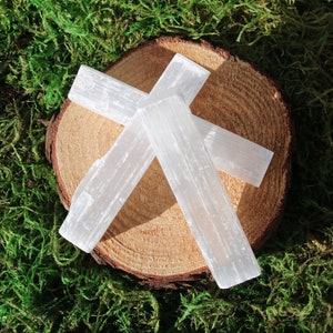 May include: Three white selenite wands arranged in a cross shape on a wooden slice. The wands are smooth and have a natural, crystalline texture.