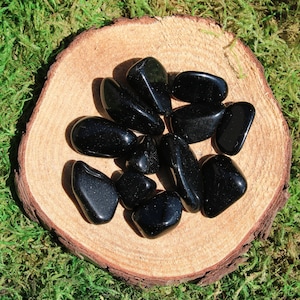 May include: A collection of black obsidian tumbled stones on a wooden surface. The stones are smooth and polished, with a deep black color.