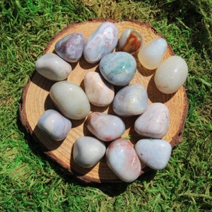 Pu&ograve; includere: Una collezione di 14 pietre di agata lisce e levigate in varie tonalit&agrave; di grigio, bianco e marrone. Le pietre sono disposte su una fetta di legno, creando un'estetica naturale e terrosa.