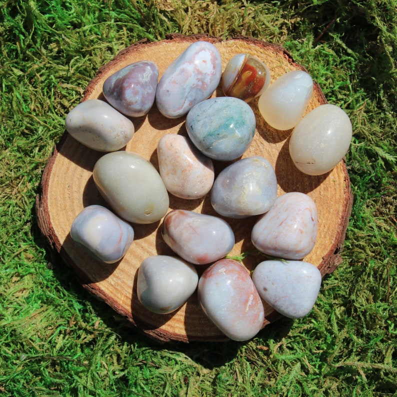 Pu&ograve; includere: Una collezione di 14 pietre di agata lisce e levigate in varie tonalit&agrave; di grigio, bianco e marrone. Le pietre sono disposte su una fetta di legno, creando un'estetica naturale e terrosa.