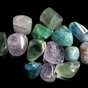 May include: A collection of tumbled stones in various shades of green, blue, and purple. The stones are smooth and polished, with a natural, organic look. Some of the stones have small labels attached to them, including one that reads "Made in Pakistan".