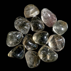 May include: A group of 11 tumbled, clear quartz stones with golden rutilated inclusions. The stones are arranged in a circle on a black background.