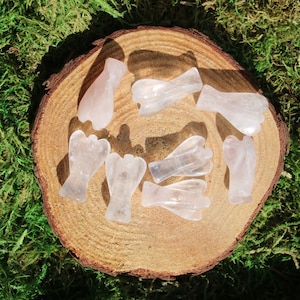May include: A collection of eight small, carved angel figurines made from clear, translucent rose quartz. The figurines are arranged on a wooden surface.