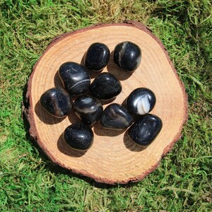 May include: A collection of ten black agate tumbled stones with white banding on a wooden slice. The stones are smooth and polished.