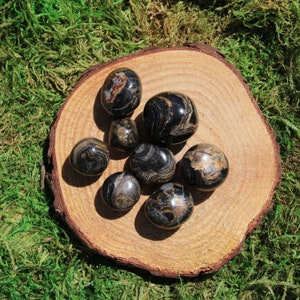 May include: A collection of eight black and gold tumbled stones on a wooden surface. The stones have a swirling pattern and are smooth to the touch.